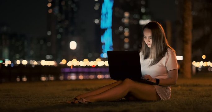 Woman developer sits in the Park at night in the city and writes code looking at the laptop screen
