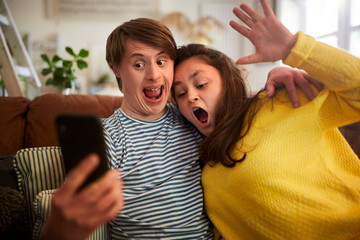 Young Downs Syndrome Couple Sitting On Sofa Using Mobile Phone To Take Selfie At Home