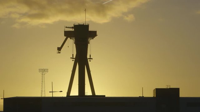 Steady, exterior, wide shot of the silhouette of large machinery and bright setting sun rays.