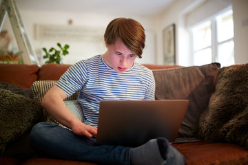 Young Downs Syndrome Man Sitting On Sofa Using Laptop At Home