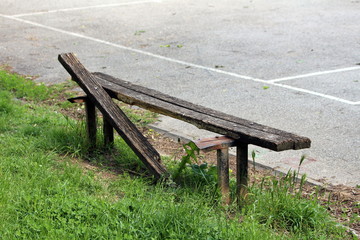 Broken wooden public bench mounted on rusted metal support next to paved playground surrounded with uncut grass on warm sunny spring day