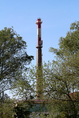 Industrial chimney with metal safety platform and multiple cell phone transmitters rising high above industrial building surrounded with tall trees and clear blue sky on warm sunny spring day