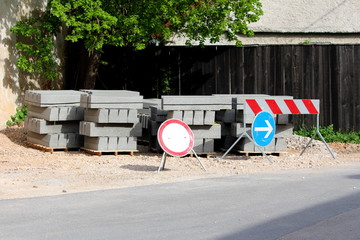 Road signs with concrete curbs on wooden pallets left as building material next to local paved road surrounded with gravel and old wooden fence on warm sunny spring day