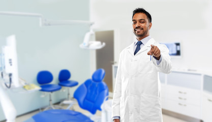 Fototapeta na wymiar medicine, dentistry and healthcare concept - smiling indian male dentist in white coat pointing to you over dental clinic office background