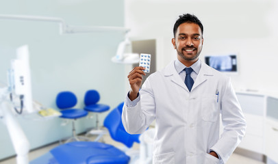 medicine, dentistry and healthcare concept - smiling indian male dentist in white coat with pills over dental clinic office background