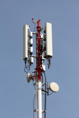Strong tall red and white metal pole with multiple cell phone transmitters and antennas mounted on each side with clear blue sky in background on warm sunny spring day