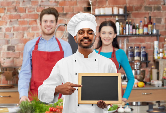 cooking class, profession and people concept - happy male indian chef in toque with blank chalkboard for menu over students background