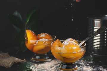 Naklejka na ściany i meble Canned peaches with syrup in a bowl on a dark background, near a tin can with a spoon, powdered sugar sprinkling on top, motion in the frame