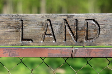 Vintage retro dilapidated sign with carved LAND on top mounted on metal pole of wire fence with...