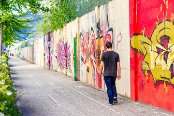 Junger Mann von hinten geht an einer Graffiti Mauer entlang an einem Radweg und Fußweg