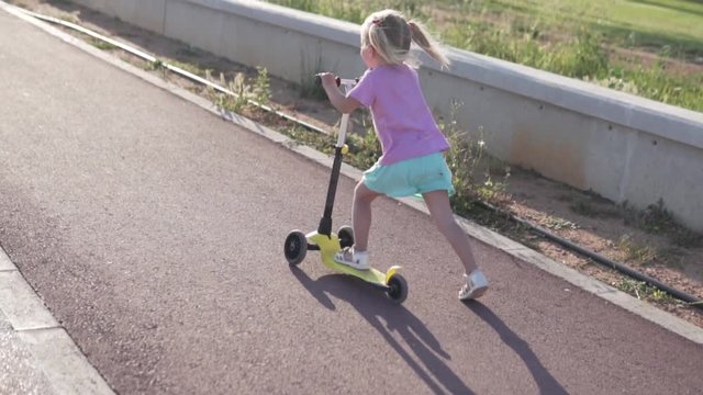 A little girl with two tails is riding a scooter in a park by the sea