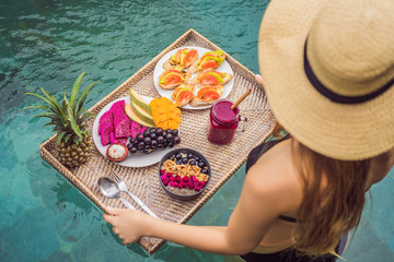 Breakfast tray in swimming pool, floating breakfast in luxury hotel. Girl relaxing in the pool drinking smoothies and eating fruit plate, smoothie bowl by the hotel pool. Exotic summer diet. Tropical
