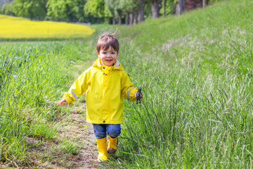 Happy little boy in yellow raincoat and muddy rubber boots running on dirt road through green grass near blooming rape seed field holding rhino in hand. Carefree childhood.
