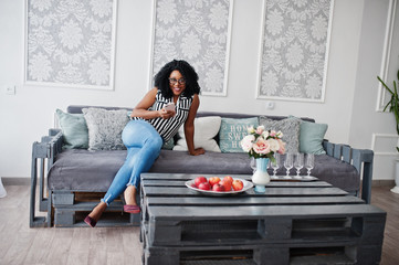 Beautiful african american woman with curly afro hair and eyeglasses, sitting on couch with mobile phone in hand.