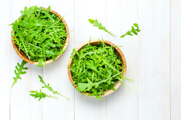 Fresh green arugula leaves on wooden bowl, rucola salad on white wooden rustic background top view with place for text. Rocket salad or arugula, healthy food, diet. Nutrition concept.