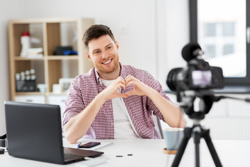 blogging, videoblog and people concept - male blogger with camera recording video and showing hand heart gesture at home office