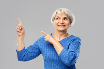 gesture and old people concept - portrait of smiling senior woman in blue sweater pointing fingers to something over grey background