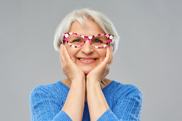 party props, photo booth and old people concept - portrait of amazed smiling senior woman in blue sweater with big glasses over grey background