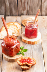 Two glass jars of strawberry and apple jam with spoons and sliced bread on wooden table.