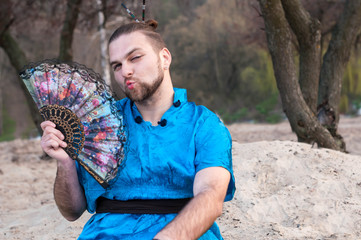 transsexual handsome man with make up, hair bun sitting on sand in blue kimono, winking, showing...