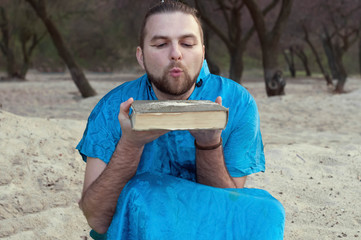 selective focus of handsome man in blue kimono blowing sand from book on beach in front of trees