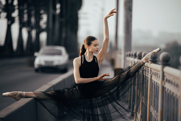 Ballerina sitting in twine pose on the bridge against the background of car and road.