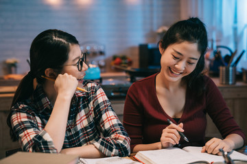 Laughing two female student learning with friend indoors at late night in dormitory kitchen. young college girls smiling having fun chatting while doing homework in midnight. women stay up late.