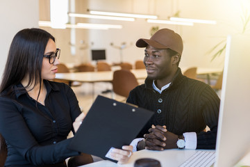 Young pretty business woman and african business man talking over clipboard in office desk