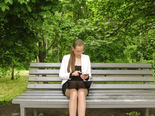 Obraz premium The girl in glasses dials the number on the screen of the smartphone sitting on a bench in a park. Working with technology outdoors concept, businesswoman with mobile and laptop