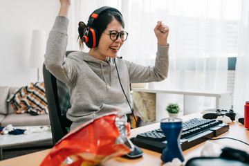 Professional gamer girl putting on headset and starts playing online video game on personal computer. Casual asian geek woman. happy female in eyeglasses winning pc game at home with yes hand gesture - obrazy, fototapety, plakaty
