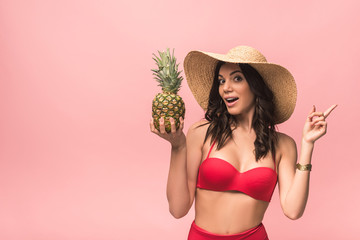 amazed girl in straw hat and bikini holding pinapple isolated on pink