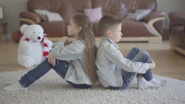 Twins boy and girl sit on the floor of living room back to back angry to each other. Brother and sister relationship. The concept of children's family love.