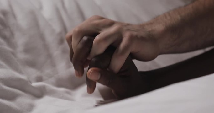 Multi-ethnic couple in love lying in bed together. Close up. Womans hand is lying on a bed, mans hand is caressing it and grabbing.