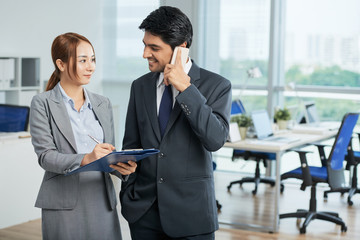 Young businessman standing in suit talking on mobile phone and smiling to young secretary while she making notes on clipboard at office