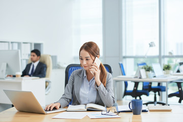 Asian young businesswoman sitting at her workplace working on laptop while talking on mobile phone with her colleague working in the background