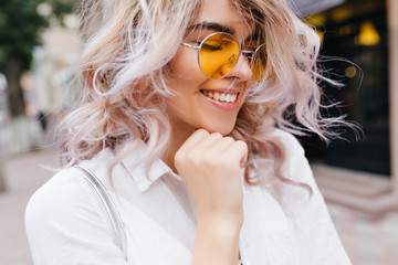 Close-up portrait of stunning young woman with blonde curls enjoying free time outside. Amazing girl in sunglasses and white jacket posing with eyes closed and smile, touching chin with fist.