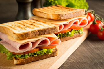 Close-up of two sandwiches with bacon, salami, prosciutto and fresh vegetables on rustic wooden cutting board. Club sandwich concept.