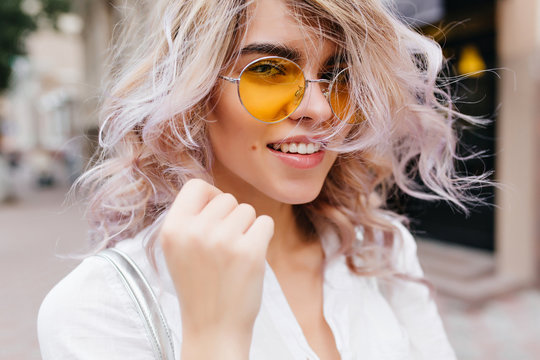 Elegant Young Woman In Trendy Accessories Posing With Smile And Holding Fist Up. Outdoor Portrait Of Lovely Blonde Girl With Romantic Hairstyle Walking Down The Street And Smiling Playfully.