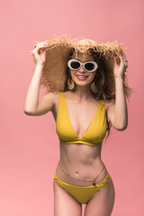 charming girl in straw hat and sunglasses smiling isolated on pink