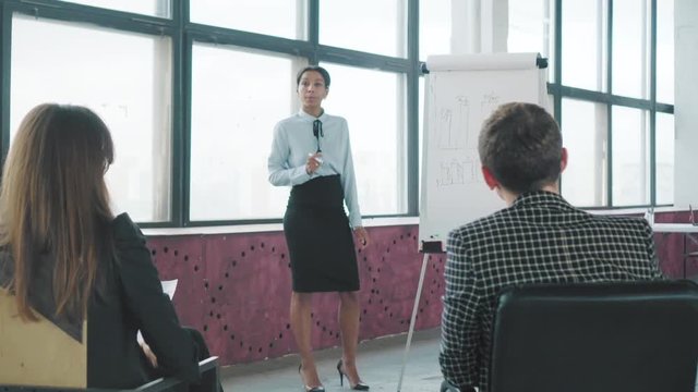 A young mixed-race female manager holds a presentation next to a flipchart and draws graphics. A colleague asks her a question. Creative office interior. Office workers