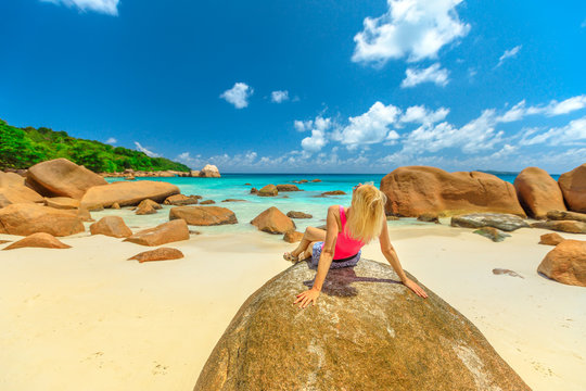 Caucasian blonde female sunbathes on a large granite boulder in popular Anse Lazio beach. Carefree woman looks turquoise waters of Indian ocean on Praslin Island, Seychelles. Sunny blue sky.