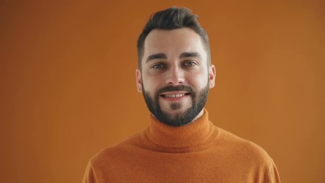 Close-up portrait of handsome young bearded man winking looking at camera and smiling against orange background. Flirtation and body language concept.