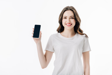 Fototapeta premium beautiful cheerful girl in white t-shirt holding smartphone with blank screen and looking at camera isolated on white