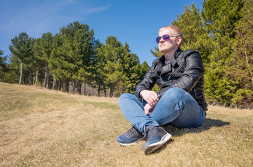 Naklejka na ściany i meble A young blond man sits in a forest glade and looks into the distance. Spring natural photography. Bright blue sky, sunny afternoon. The concept of outdoor recreation, tranquility, reflection.