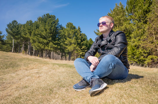 A young blond man sits in a forest glade and looks into the distance. Spring natural photography. Bright blue sky, sunny afternoon. The concept of outdoor recreation, tranquility, reflection.