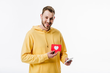 cheerful young man using smartphone while holding red paper cut card with heart symbol isolated on white