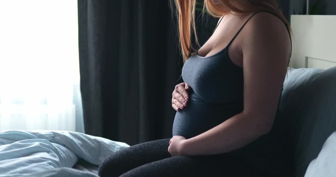 Young pregnant woman sitting on bed and touching her belly