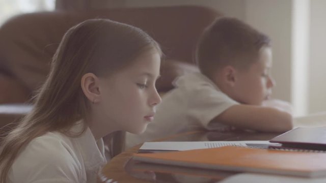 Sister and brother sit on the table and making homework from school. Girl fall asleep on the table. Boy still reading a book.