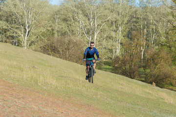 Obraz premium Cyclist in shorts and jersey on a modern carbon hardtail bike with an air suspension fork rides off-road on green hills near the forest 