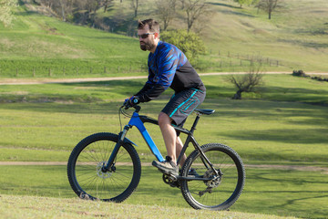 Obraz premium Cyclist in shorts and jersey on a modern carbon hardtail bike with an air suspension fork rides off-road on green hills near the forest 